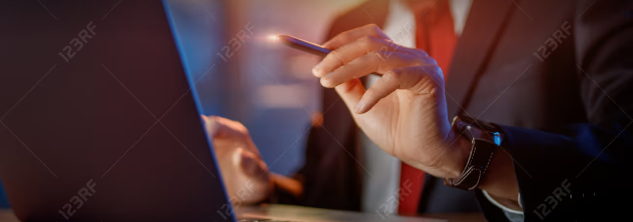 Close-up of a hand holding a stylus near an open laptop, with a colleague in a suit visible in the background.